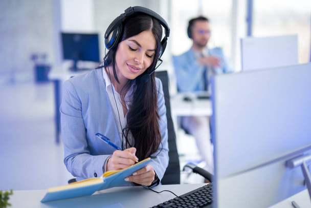 Γυναίκα τηλεφωνήτρια φορώντας ακουστικά κρατώντας σημειώσεις κατά την εργασία σε υπολογιστή, επικοινωνώντας με τους πελάτες και επιλύοντας τα προβλήματά τους. - Φωτογραφία, εικόνα