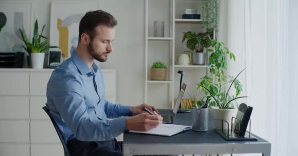Lockdown-Aufnahme eines jungen Geschäftsmannes, der Strategie in Tagebuch schreibt, während er Laptop am Schreibtisch im Büro benutzt - Filmmaterial, Video