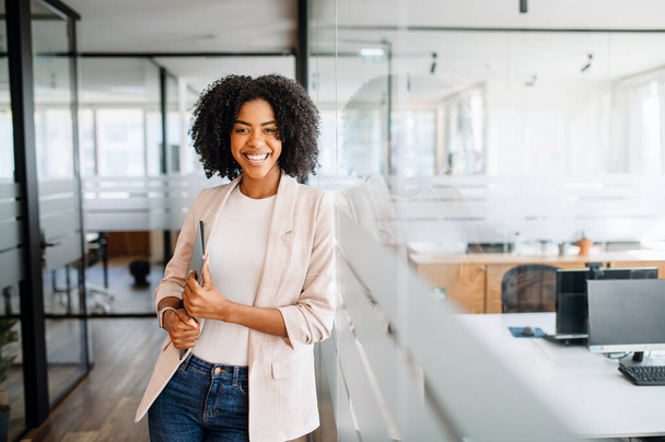A confident young woman with a bright smile stands by a glass partition, holding the laptop in her hand, embodying professionalism and readiness. The concept of career advancement - Foto, imagen