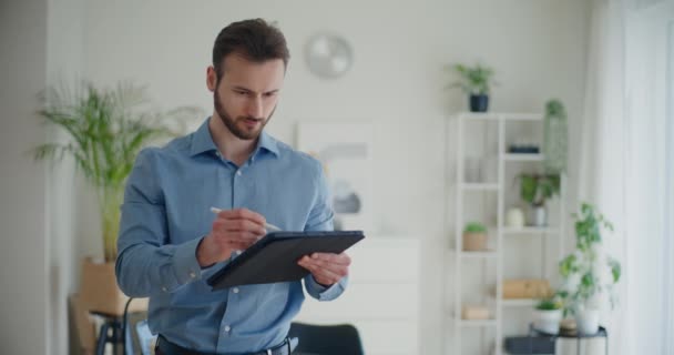 Blocco colpo di fiducioso giovane imprenditore maschile utilizzando tablet digitale con stilo mentre in piedi nello spazio di lavoro aziendale - Filmati, video