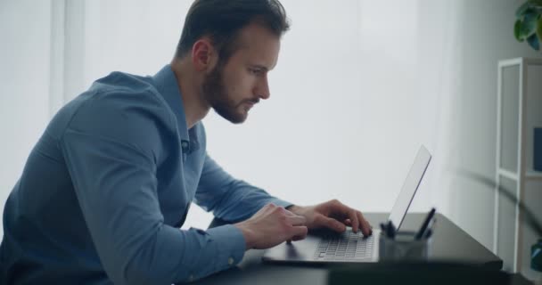 Πλευρική άποψη του νεαρού γενειοφόρου επιχειρηματία που χρησιμοποιεί φορητό υπολογιστή κάθεται στο γραφείο εργασίας στο εταιρικό γραφείο - Πλάνα, βίντεο
