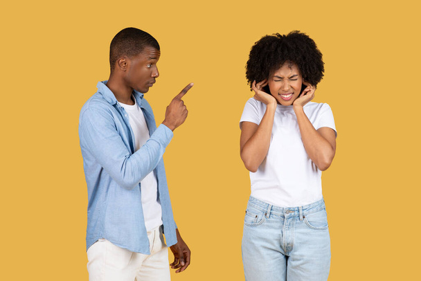 Eine aufgebrachte schwarze junge Frau verdeckt ihre Ohren, während ein junger wütender Mann auf sie zeigt, beide stehen vor einem schlichten senfgelben Hintergrund, einem Studio. Beziehungsprobleme - Foto, Bild
