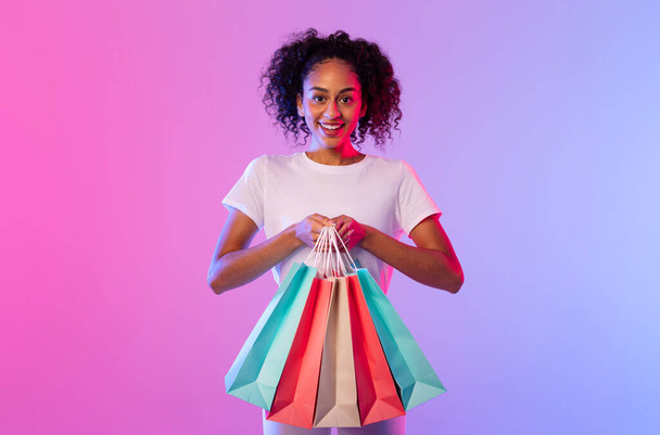 Kıvırcık saçlı parlak siyah kadın alışveriş torbaları tutuyor, başarılı alışveriş çılgınlığı sergiliyor, çarpıcı pembe ve mavi gradyan plana karşı, tüketici sevinci ve perakende terapisi yayıyor. - Fotoğraf, Görsel