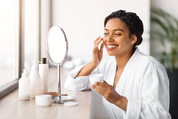 Femme noire souriante en peignoir appliquant de la crème sur sa joue, belle femme afro-américaine bénéficiant d'une routine de soins de la peau, assise devant un miroir de maquillage avec des produits de beauté sur la table - Photo, image