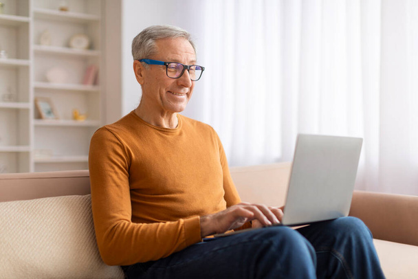 ラップ,ウェブサーフィン,オンラインニュースを読むラップ上のラップトップコンピュータとソファーに座っているポジティブな祖父. 高齢者は自宅からオンラインで働いています. 高齢者のためのリモートジョブ - 写真・画像