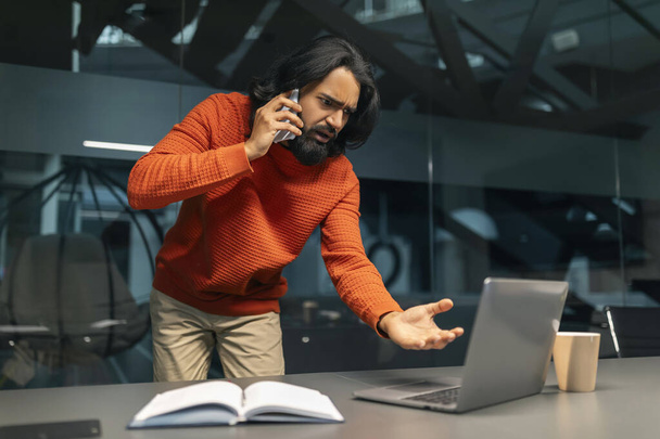 Stresszes millenniumi indiai menedzser vagy üzletember harcol telefonon asszisztensével, nézi laptop számítógép képernyő, gesztikuláló, sötét modern irodai belső tér, másolás tér - Fotó, kép