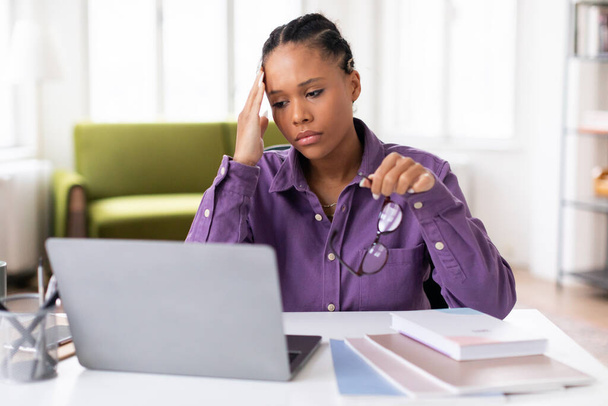 Zaniepokojona czarna studentka nosząca purpurową koszulę, wykazująca oznaki stresu podczas nauki w laptopie, trzymająca okulary i dotykająca głowy, siedząca za biurkiem w domu - Zdjęcie, obraz