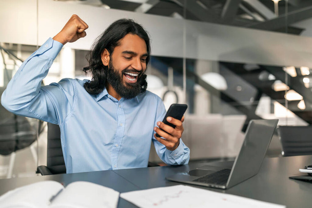 Gestionnaire de gars indien excité regardant l'écran du smartphone, geste levant la main et criant, avoir de bonnes nouvelles, célébrer le succès de l'entreprise, intérieur de bureau moderne, espace de copie - Photo, image
