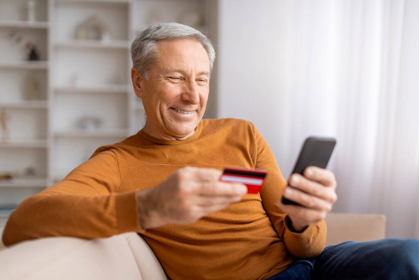 Χαμογελώντας παππούς κρατώντας κόκκινο πλαστικό πιστωτική κάρτα και smartphone, κάθεται στον καναπέ στο σπίτι, πληρώνουν για αγαθά και υπηρεσίες σε απευθείας σύνδεση, αντίγραφο χώρου. Εύκολα ψώνια, εικονική τραπεζική - Φωτογραφία, εικόνα