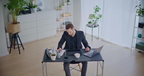 Pewny siebie młody biznesmen piszący strategię na papierze podczas korzystania z laptopa siedzącego przy biurku w korporacyjnej przestrzeni roboczej - Materiał filmowy, wideo