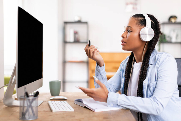 Örgülü siyah kadın modern kulaklıklar takıyor, boş bilgisayar ekranına bakarken düşünceli bir şekilde işaret ediyor, çevrimiçi ders veriyor. - Fotoğraf, Görsel
