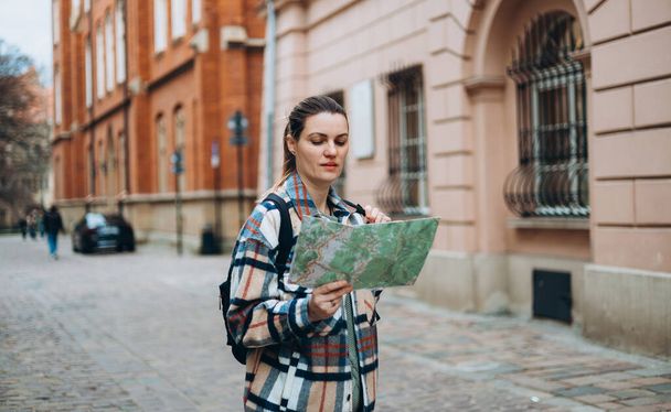Şehir caddesinde yürüyen haritalı neşeli genç bir kadın, şehir yaşam tarzı konsepti. Gezgin. Sırt çantalı çekici bayan turist yeni bir şehri keşfediyor. - Fotoğraf, Görsel