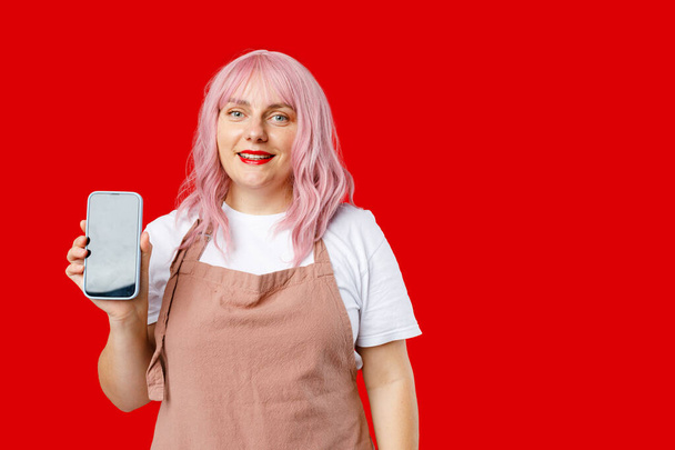 Отличное мобильное предложение. Возбужденная женщина 30-х годов с розовыми волосами в фартуке, держащая смартфон в руке, эмоционально реагирует на новое приложение. Женщина позирует на красном фоне - Фото, изображение