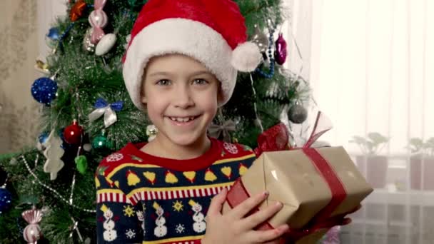 Una niña sonriente con un sombrero rojo de Santa Claus sacude alegremente un regalo de Navidad. La alegría de la fiesta encarnada en la deliciosa apertura de un regalo de niños. Primer plano. - Metraje, vídeo