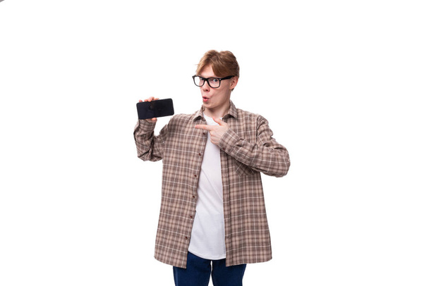νεαρός κομψός άνδρας με κόκκινα μαλλιά ντυμένος με καλοκαιρινό πουκάμισο δείχνει ένα τηλέφωνο με ένα mockup. - Φωτογραφία, εικόνα