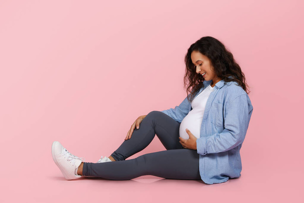 Happy young pregnant woman sitting on floor isolated on pink studio background, embracing her big tummy and smiling, enjoying pregnancy, waiting for baby to come, copy space - Photo, Image
