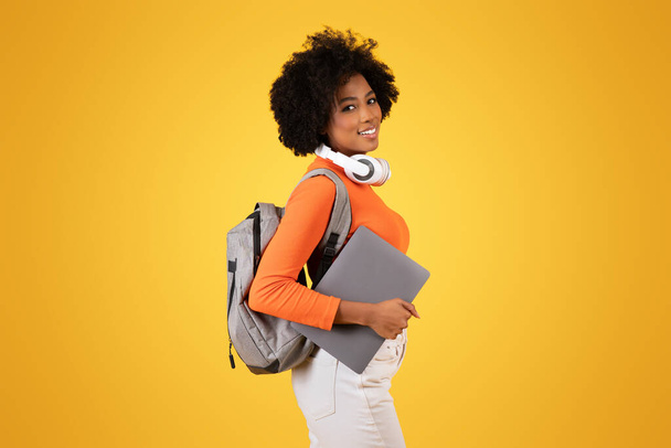 Счастливая афроамериканка с ноутбуком и наушниками на шее с улыбкой оглядывается назад, надевая рюкзак и оранжевую рубашку. - Фото, изображение