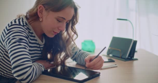 Впевнена приваблива молода жінка використовує стилус для написання на цифровому планшеті, сидячи за столом вдома - Кадри, відео