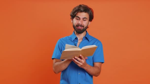 Young excited middle eastern man reading funny interesting fairytale story book, laughing, leisure hobby, knowledge wisdom, education learning study, wow. Arab guy isolated on orange studio background - Footage, Video