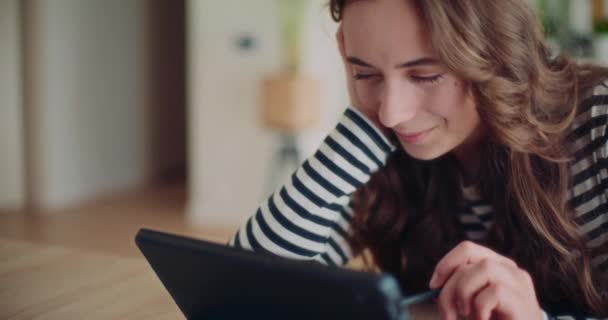 Улыбающаяся юная брюнетка, прокручивающая социальные сети через цифровой планшет, опираясь на стол дома - Кадры, видео