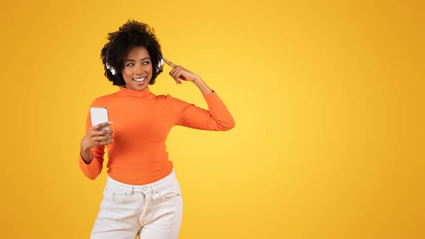 Χαμογελώντας millennial Αφροαμερικανή γυναίκα με σγουρά μαλλιά ακούγοντας μουσική σε ασύρματα ακουστικά και κρατώντας ένα smartphone σε ένα πορτοκαλί ζιβάγκο σε κίτρινο φόντο - Φωτογραφία, εικόνα