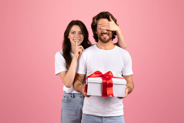 Verrassende blanke man met baard in een wit shirt houdt onbewust een geschenk met een rood lint vast als een lachende vrouw met een vinger op haar lippen in het geheim de verrassing onthult op een roze achtergrond - Foto, afbeelding
