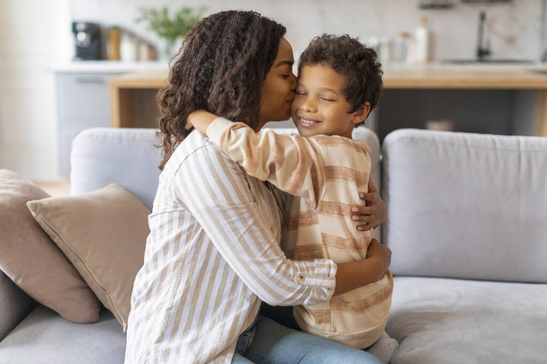 Mamá negra besándose y abrazando a su hijo preadolescente mientras se sientan juntos en el sofá, madre afroamericana feliz y niño varón que se une en casa, mamá y niño disfrutando del tiempo con los demás - Foto, Imagen