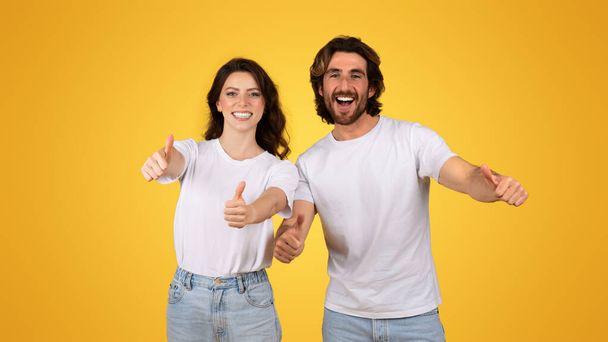 Strahlendes europäisches Paar, das Daumen hoch gibt, Zustimmung und Glück zeigt, in weißen T-Shirts und Jeans gekleidet, mit fröhlichem Gesichtsausdruck auf fröhlich gelbem Hintergrund, Studio - Foto, Bild