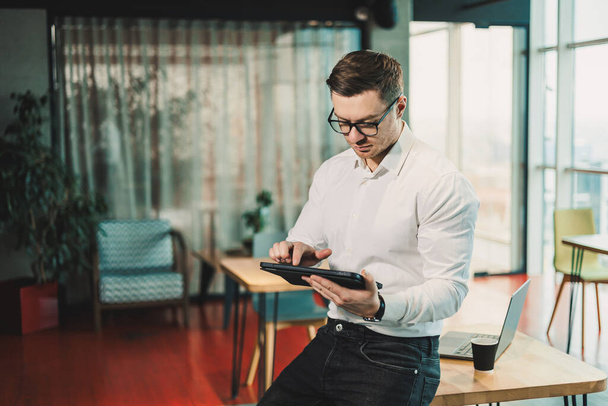 Ein junger Mann im weißen Hemd steht in einem geräumigen modernen Büro und arbeitet an einem Tablet. Moderne Büroangestellte. Ein Geschäftsmann mit Brille arbeitet online. - Foto, Bild
