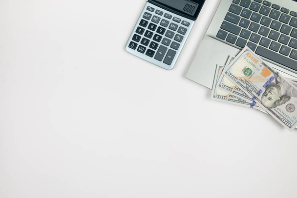 Κάτοψη του laptop και αριθμομηχανή με δολάρια σε λευκό φόντο με αντίγραφο χώρου. - Φωτογραφία, εικόνα