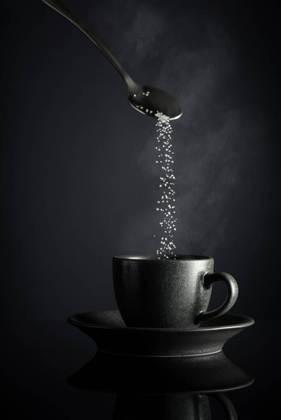 黒い反射の背景にあるコーヒーのブラックカップ. 砂糖は一杯のコーヒーに注ぎ込まれています. コピースペース. - 写真・画像