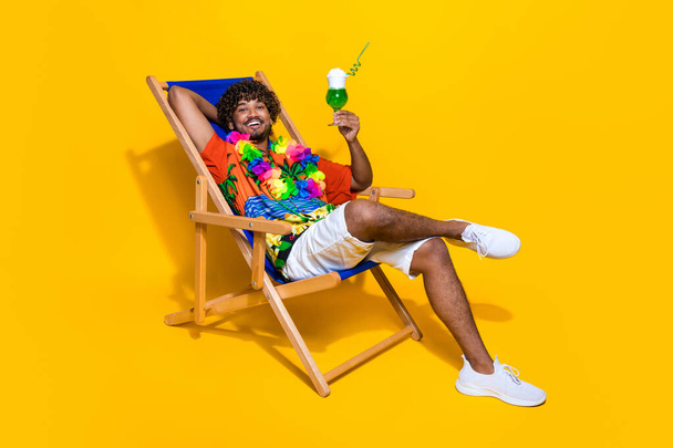 Full length photo of relaxed guy wear hawaii shirt drink green cocktail sit on chair on vacation isolated on yellow color background. - Valokuva, kuva