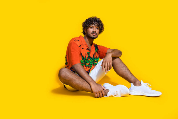 Full size photo of confident serious man dressed print shirt white shorts shoes sitting on floor isolated on yellow color background. - Fotó, kép