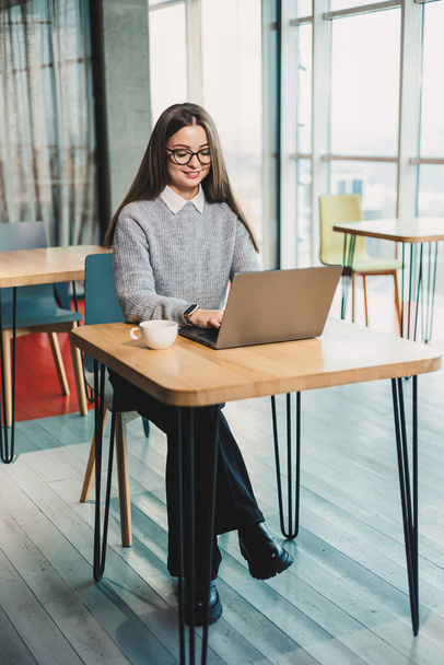 Милая деловая женщина, сидящая за столом в современном рабочем пространстве и работающая над ноутбуком. Рабочий процесс женщины в офисе - Фото, изображение