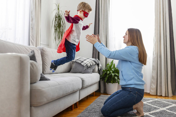 Brincadeira alegre entre uma mãe e seu filho fingindo voar como um super-herói em uma sala de estar. - Foto, Imagem