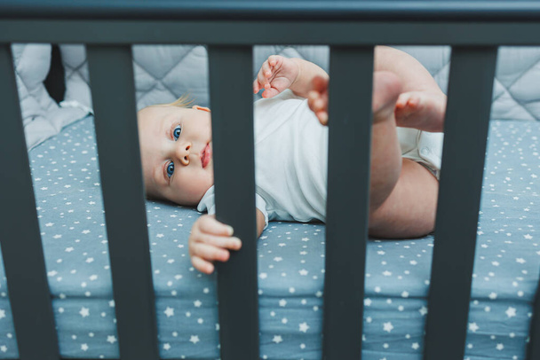 The baby looks through the sides of the crib into the room. A small newborn boy lies in a crib for children. A place for children to sleep - Photo, Image