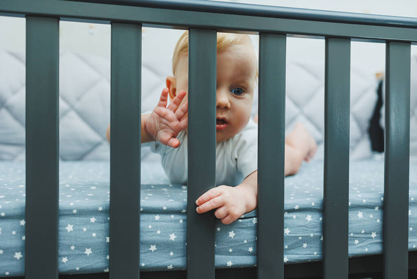 Ребенок смотрит через край кроватки в комнату. Маленький новорожденный мальчик лежит в детской кроватке. Место для сна детей - Фото, изображение