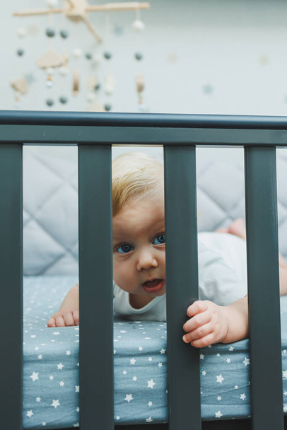 Το μωρό κοιτάζει μέσα από τις πλευρές της κούνιας στο δωμάτιο. Ένα μικρό νεογέννητο αγόρι βρίσκεται σε μια κούνια για παιδιά. Ένα μέρος για να κοιμούνται τα παιδιά - Φωτογραφία, εικόνα