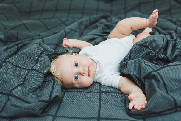 Ein kleines Kind liegt zu Hause auf einem großen Bett. Porträt eines fünf Monate alten Babys, das auf einem grauen Bett liegt. Fröhliches Glückskind. - Foto, Bild