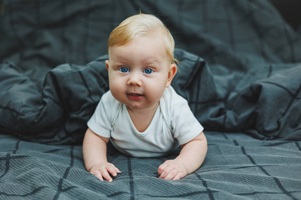 Ένα μικρό παιδί είναι ξαπλωμένο σε ένα μεγάλο κρεβάτι στο σπίτι. Πορτρέτο ενός μωρού πέντε μηνών ξαπλωμένου σε ένα γκρι κρεβάτι. Χαρούμενο ευτυχισμένο παιδί.. - Φωτογραφία, εικόνα