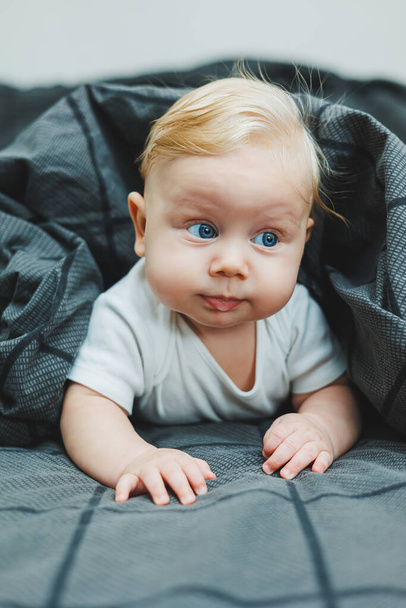 Een klein kind ligt thuis op een groot bed. Portret van een vijf maanden oude baby liggend op een grijs bed. Vrolijk gelukkig kind. - Foto, afbeelding