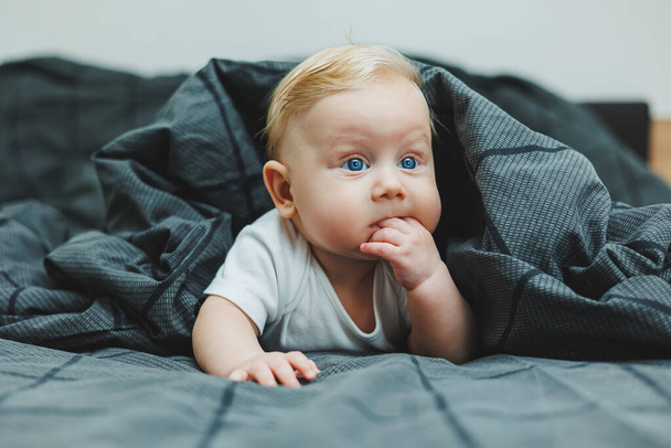 Ένα χαρούμενο αγοράκι με όμορφα μεγάλα μάτια είναι ξαπλωμένο στο κρεβάτι στο σπίτι με ένα λευκό κοστούμι σώματος σε γκρι κρεβάτι. Το παιδί είναι ξαπλωμένο στο κρεβάτι στο σπίτι - Φωτογραφία, εικόνα