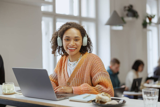Una mujer que usa auriculares está sentada en una mesa, usando una computadora portátil. Ella sonríe mientras trabaja en su computadora personal - Foto, imagen