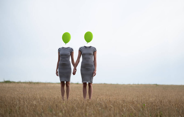δύο χαρούμενες σέξι νέες γυναίκες, σε μαύρο και άσπρο ριγέ φόρεμα, τους φίλους, κρατώντας τα χέρια, τα κεφάλια που αντικαθίστανται από πράσινα μπαλόνια σταθεί χέρι-χέρι στο συγκομισμένο χωράφι καλαμποκιού, αντίγραφο χώρο - Φωτογραφία, εικόνα