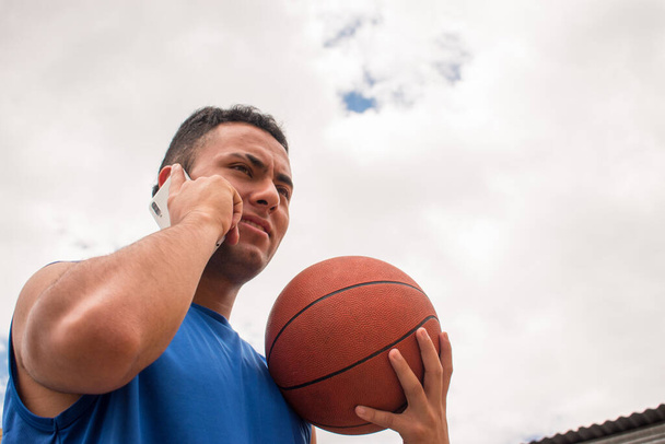 Bajo ángulo de hombre sosteniendo pelota de baloncesto y hablando de teléfono en la cancha en verano  - Foto, Imagen