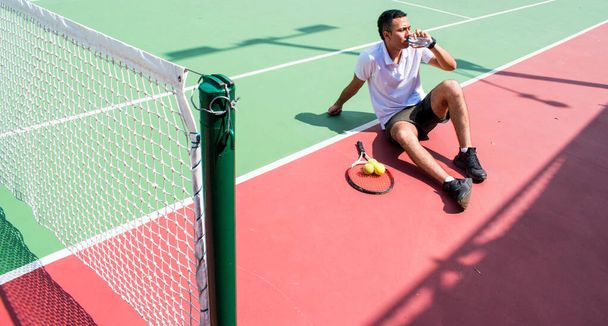 Jonge latn man tennisser rusten en drinken van water op de tennisbaan in zonnige hete dag in de zomer - Foto, afbeelding