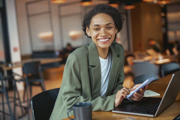 Una mujer en un traje de trabajador de cuello blanco está sentada en una mesa con una computadora portátil y un teléfono celular, sonriendo mientras trabaja en la computadora - Foto, imagen