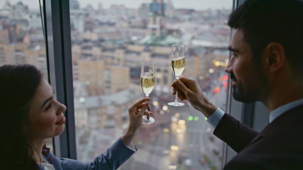 Koledzy świętujący osiągnięcie z szampanem stojącym przy oknie panoramicznym biura z bliska. Dwóch udanych współpracowników pijących wino, radujących się z zawarcia umowy biznesowej. Koncepcja partnerstwa. - Zdjęcie, obraz