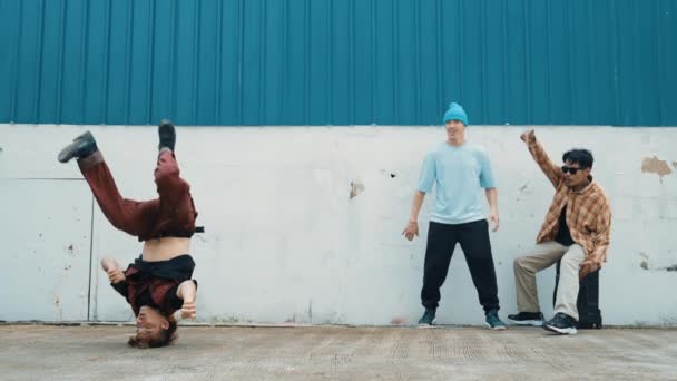 Mladý asijský tanečník tančí v hip hop stylu s multikulturní přátelé jásají za zdí. Atraktivní pohledný muž natahuje ruce na ulici s modrým pozadím. Outdoor sport 2024. hiphop. - Záběry, video