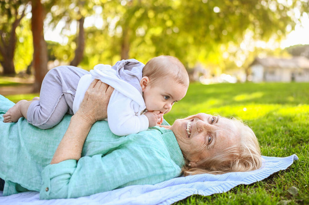 Beautiful happy smiling senior elderly woman lying on grass with cute little boy. Бабушка и внук веселятся вместе в тропическом летнем парке. Семейная концепция - Фото, изображение